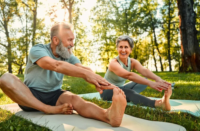 flexible exercises for body sporty man and woman with grey hair stretching on yoga mats with hands to one leg during outdoors workout happy married couple with bare feet warming up together at park