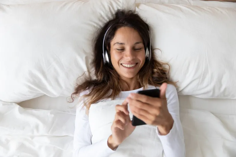 top view close up smiling woman wearing headphones enjoying music, holding phone, resting on soft pillow in bedroom in morning, happy beautiful girl listening to favorite popular song, lying in bed