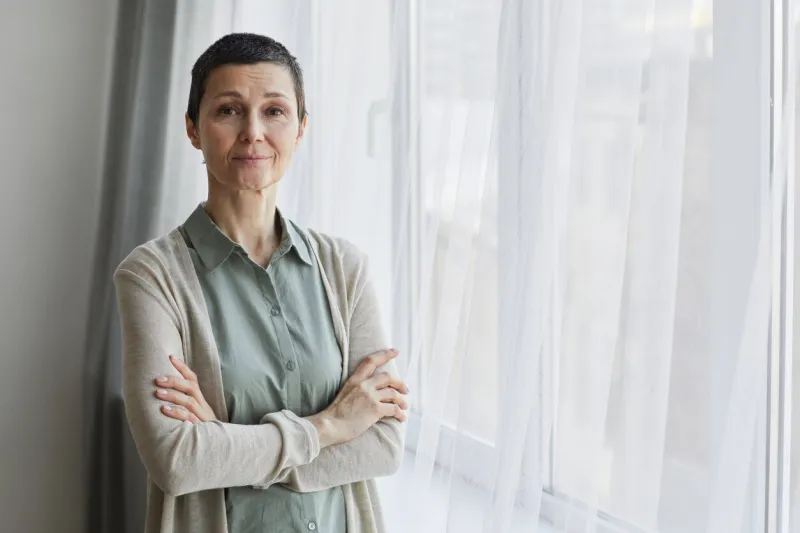 waist up portrait of mature woman looking at camera while standing by window with arms crossed, copy space