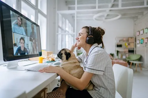 photo of a smiling young woman having a video call, accompanied by her dog, the daily routine of a young woman working from home