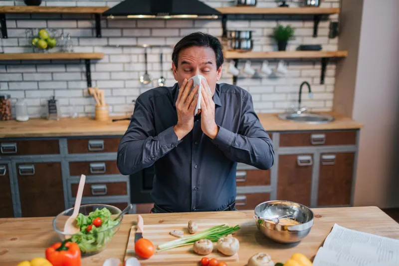 sick adult man sneezing to white napkin he stand at table in kitchen desk full of colorful healthy vegetables and spices