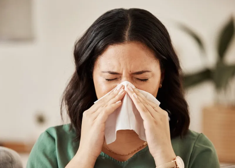 blowing nose, tissue and woman in a living room with flu, cold and hay fever, crisis or viral infection in her home sneezing, allergies and person with virus or coughing illness in a lounge in house