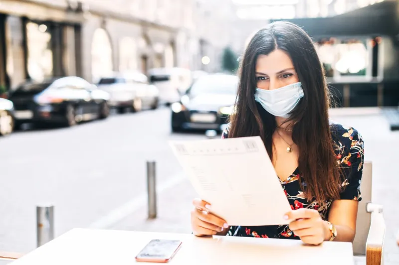 dinner in the summer cafe after quarantine relief young woman with a medical mask on the face sits in cafe with a menu in hands and going to make a order