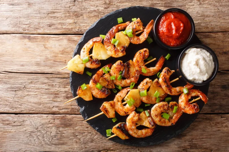 fresh kebabs made from shrimps and pineapple slices decorated with green onions served with sauces close-up on a slate plate on a wooden table horizontal top view from above