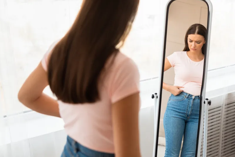 unhappy woman gaining excess weight touching drooping belly standing at mirror at home unsuccessful diet selective focus