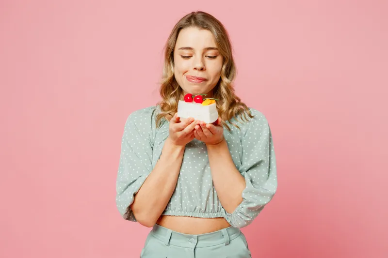 young smiling satisfied fun happy woman she wear casual clothes look at tasty piece of seet cake dessert, lick lips isolated on plain pastel light pink background studio portrait lifestyle concept