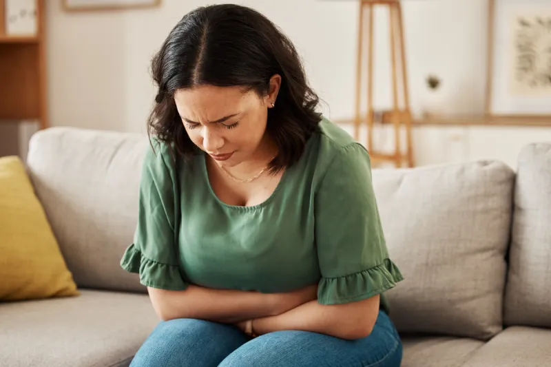 stress, stomach pain and woman on a sofa with menstruation, gas or constipation, pms or nausea at home gut health, anxiety and lady with tummy ache in living room from ibs, bloated or endometriosis