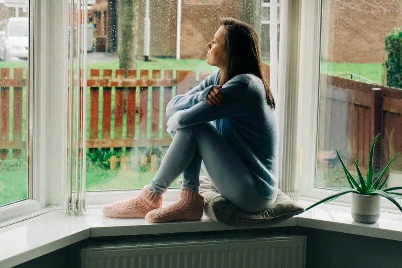 thoughtful sad woman sitting on a windowsill lonely young woman feeling depressed and unhappy mental health and seasonal disorder due to rainy cold weather