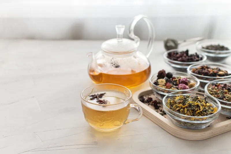 glass cup with hot tea and a kettle on a bright table by the window tray with different varieties of herbal tea on a white background delicious and healthy drink to strengthen immunity space for text