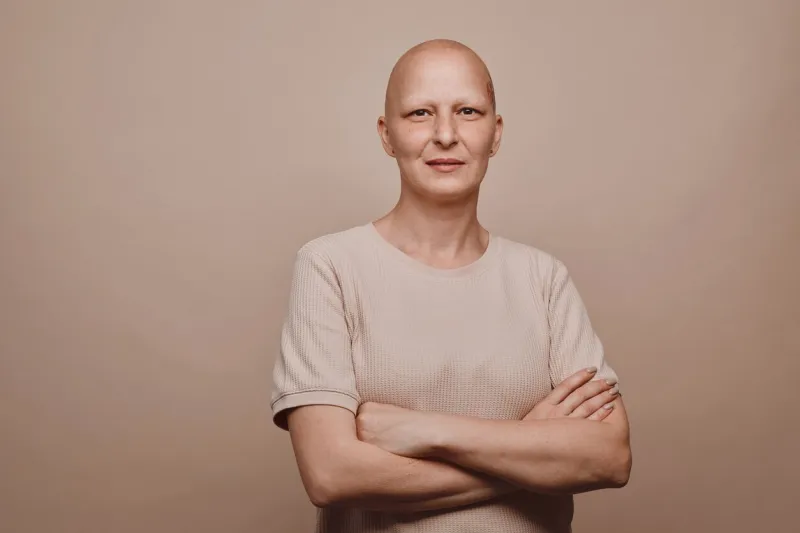 warm-toned waist up portrait of confident bald woman looking at camera while posing against minimal beige background in studio, alopecia and cancer awareness, copy space