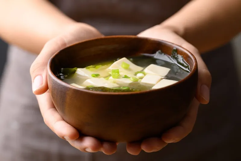 miso soup with tofu, wakame seaweed and spring onion in wooden bowl holding by hand, traditional japanese soup
