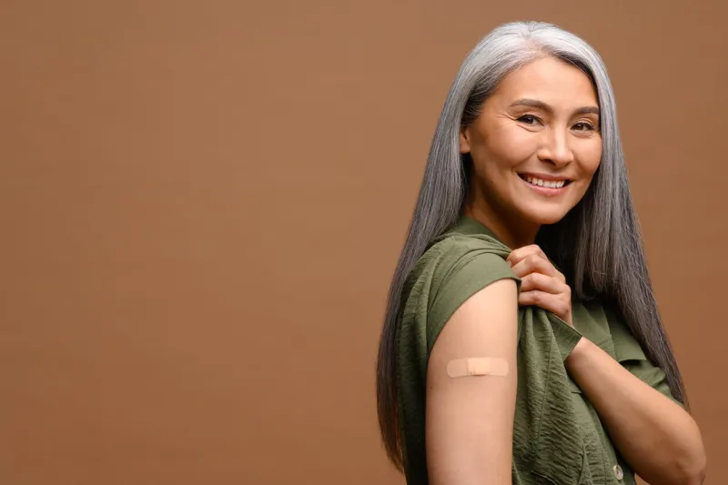 smiling senior grey-haired woman with a medical patch after vaccination isolated on brown background, protecting hand with bandage after injection healthcare and medicine concept