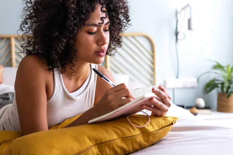 pensive young multiracial latina woman lying down on bed with serious expression writing on journal in cozy bedroom lifestyle concept