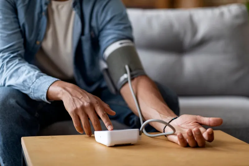 cropped shot of young black man checking blood pressure with upper arm monitor while sitting on couch at home, unrecognizable african american male measuring arterial tension, closeup