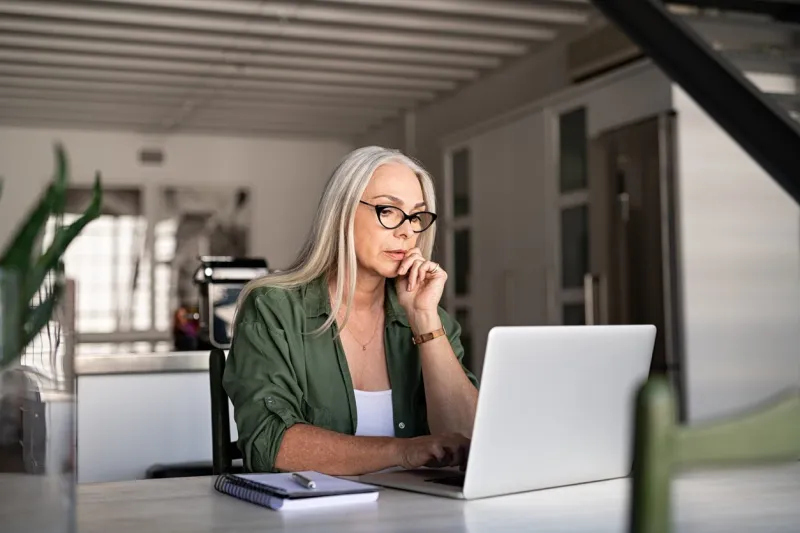 focused old woman with white hair at home using laptop senior stylish entrepreneur with notebook and pen wearing eyeglasses working on computer at home serious woman analyzing and managing domestic bills and home finance