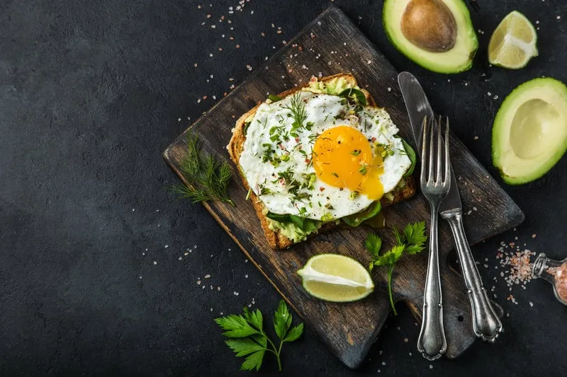 toast with avocado, spinach and fried egg on wooden cutting board, black background, top view