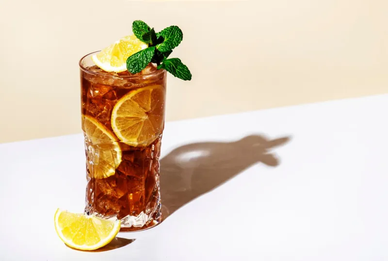 long island ice tea cocktail drink with vodka, rum, tequila, gin, liquor, lemon juice, cola and ice with lemon slice and mint in highball glass beige background, hard light, minimalism