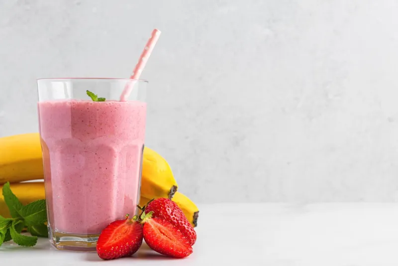 strawberry and banana smoothie or milkshake in a glass with straw, fresh berries and mint on white background summer refreshing drink