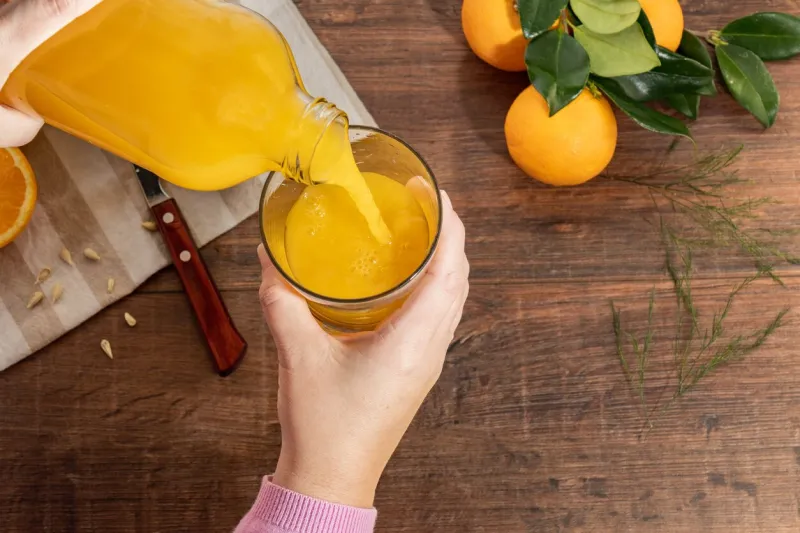 a person pouring orange juice into a glass cup in the background you can see whole oranges and a knife resting on a wooden table