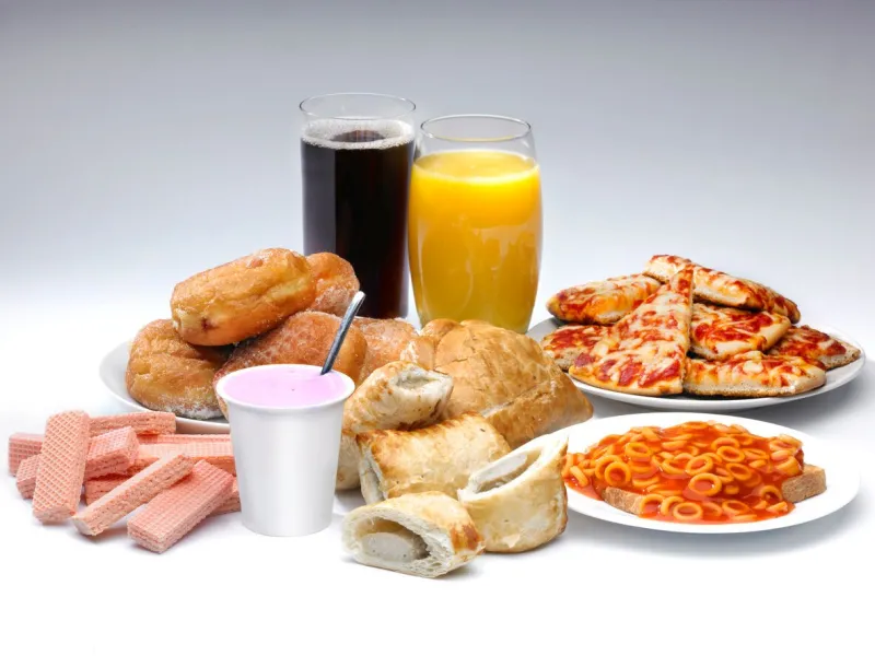 a selection of processed food and drink on a white background including pizza, cola, buscuits and sausage rolls