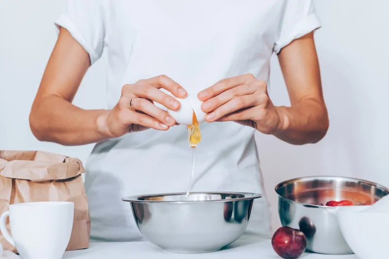 woman wearing white t-shirt breaking egg while cooking pie female baker standing at table with metal bowl, fresh fruits, sack of flour, cup and preparing dough for plum cake