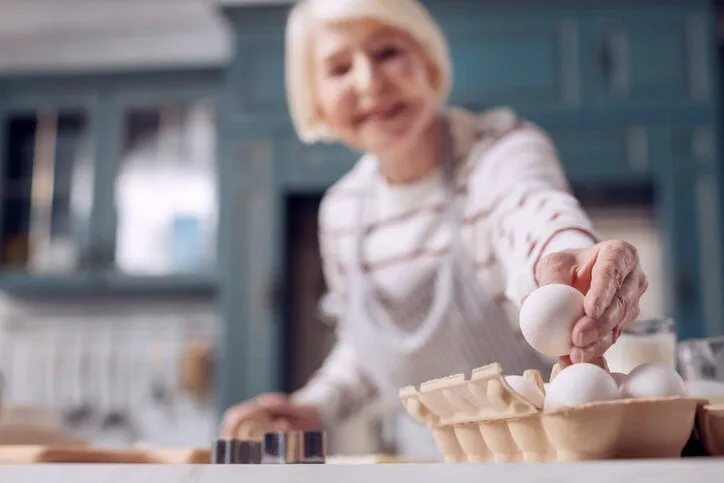 important ingredient the focus being on the hand of the pleasant elderly female taking an egg from an egg carton while making dough
