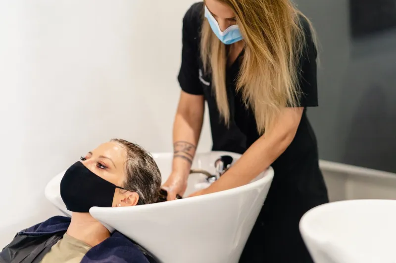 female hairdresser washing a client's head in a salon, protected by a mask against covid-19