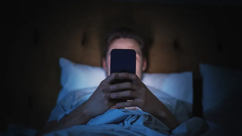 anonymous man uses smartphone in bed at home at night handsome guy browsing social media, reading news, doing online shopping late at night focus on hand holding mobile phone covering face