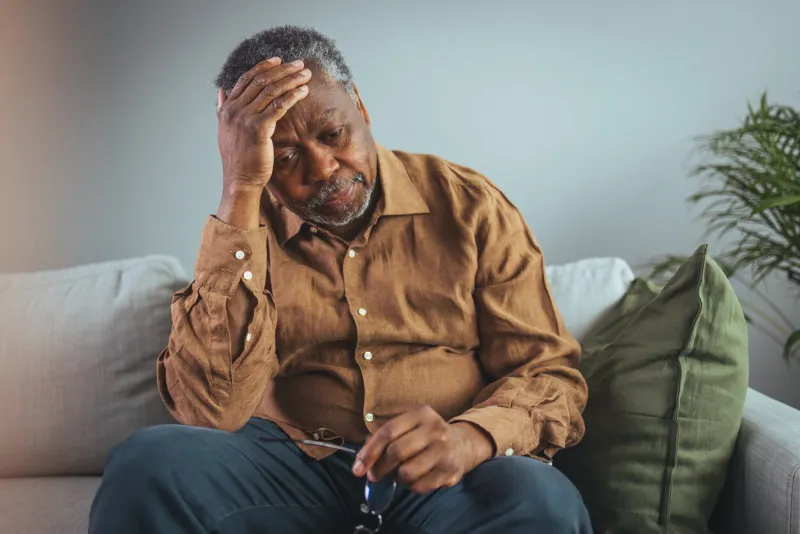 a senior black male grips his glasses, showing signs of distress while seated alone on a sofa, reflecting potential health concerns in a home setting