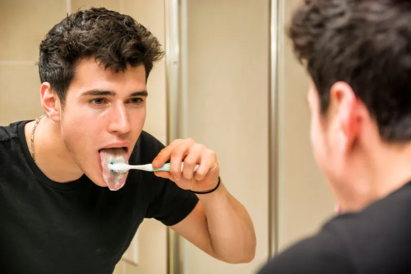 headshot of attractive young man brushing teeth and cleaning tongue with toothbrush, looking at himself in mirror