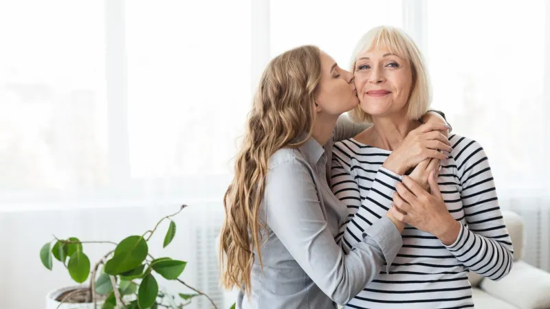 young daughter kissing senior mother on the cheek, standing against window at home, copy space