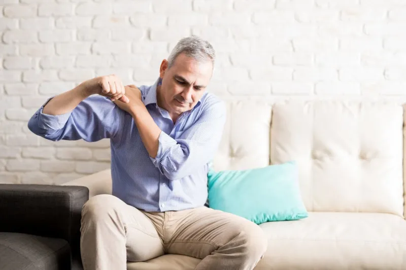 middle aged man sitting on the sofa and holding painful shoulder with another hand