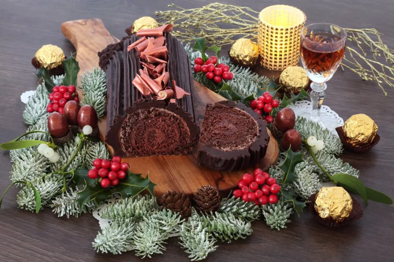yule chocolate decorated log cake for the christmas season with winter flora, alcoholic drink and foil wrapped chocolates on rustic oak wood background