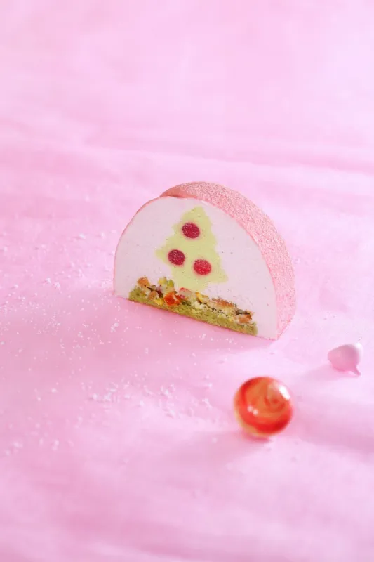 contemporary yule log cake with pistachio sponge, candied fruits, pistachio mousse, red currant jelly, cream cheese mousse and white chocolate velvet coating, decorated with white chocolate elements, on light pink background