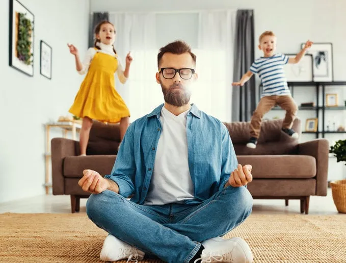 tranquil young man in casual wear sitting on floor in lotus position and meditating while kids having fun and jumping on sofa at home