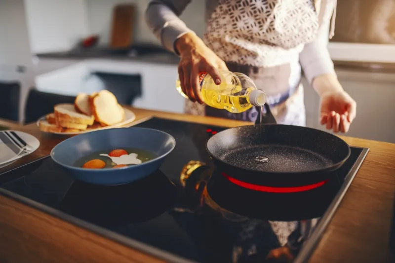 cropped picture of woman in apron pouring oil in frying pan while standing next to stove breakfast preparation concept