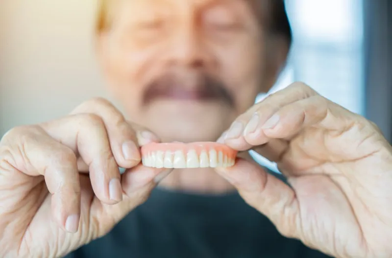 grey haired senior man holding orthodontic prosthesis denture with a happy face standing and smiling with a confident selective focus on hands