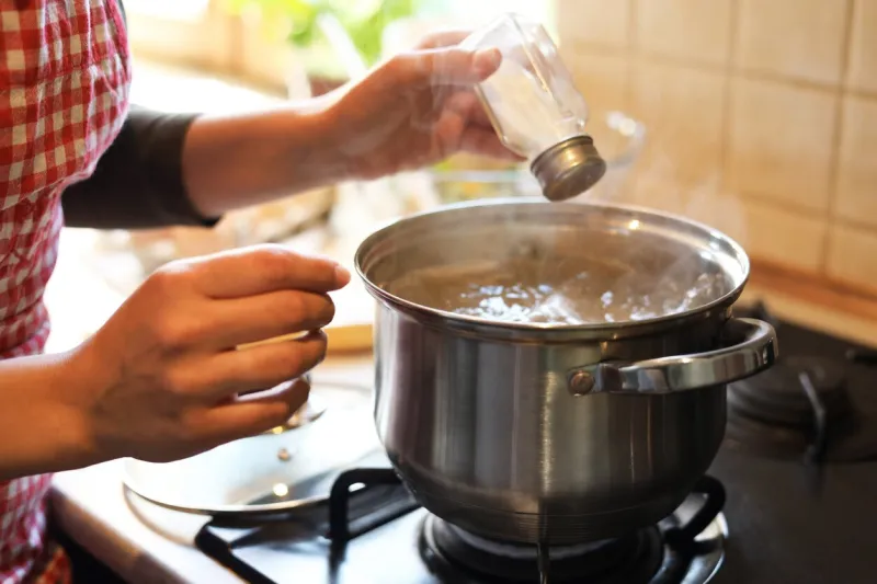 woman adding salt to boiling water in pot on stove indoors, closeup