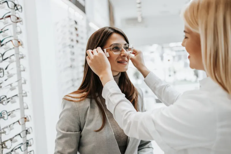 beautiful and fashionable woman choosing eyeglasses frame in modern optical store female seller specialist helps her to make right decision