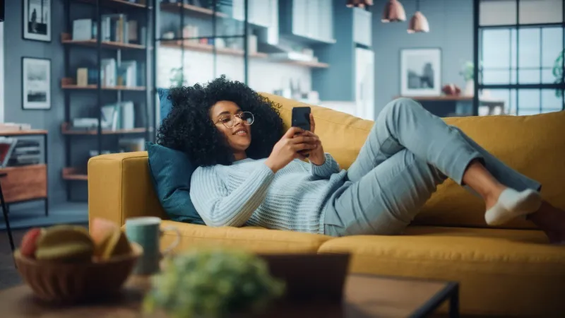 happy beautiful latina female using smartphone in cozy living room at home female resting on comfortable sofa she's browsing the internet and checking videos on social networks and having fun