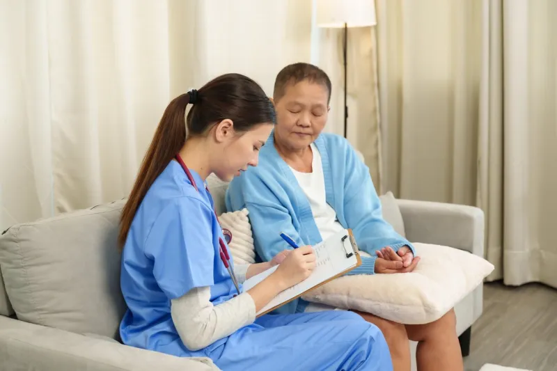 young nurse writing on clipboard while consulting elderly woman sitting on couch during recovery focus on documenting patients condition and providing personalized care in home setting