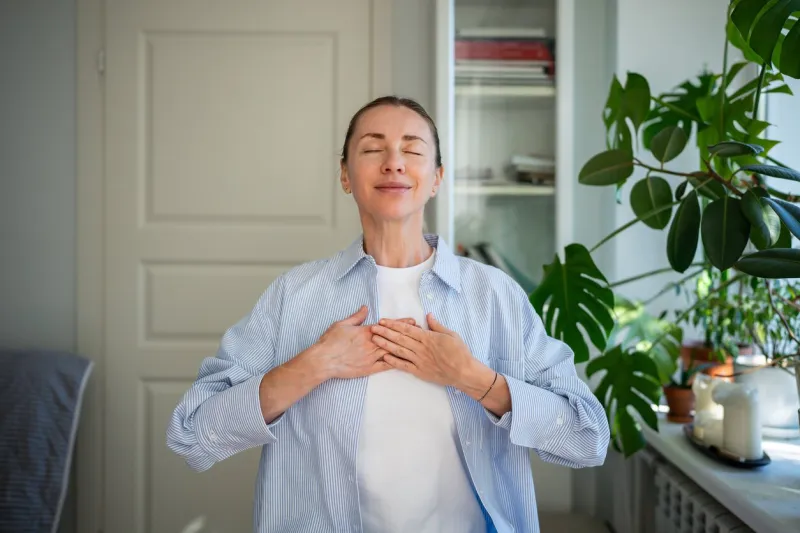 relaxed smiling woman meditates with hands on chest, practicing breathing exercises at home mindfulness practice, focus on breath, self care routine, stress relief, reflection and holistic wellness