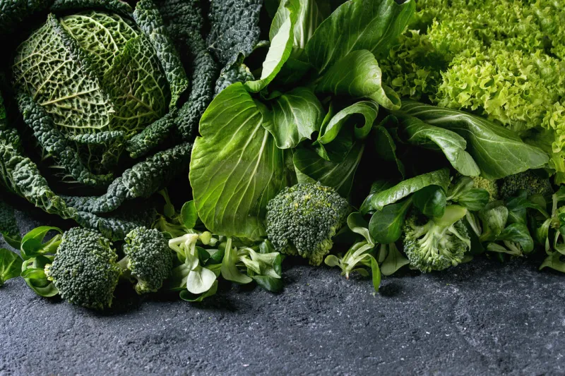 variety of raw green vegetables salads, lettuce, bok choy, corn, broccoli, savoy cabbage as frame over black stone texture background space for text