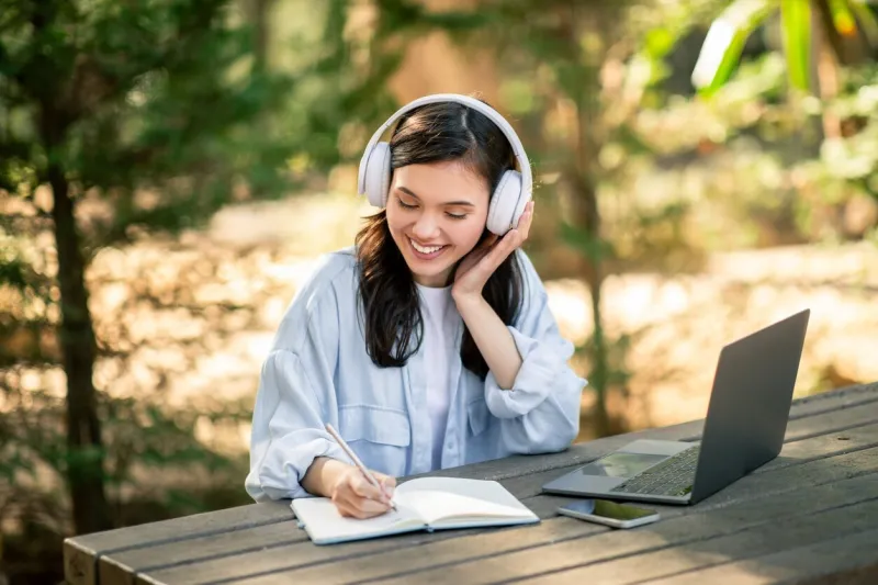 cheerful woman enjoys music while writing in a notebook, laptop on table outdoors at public park student have online lesson