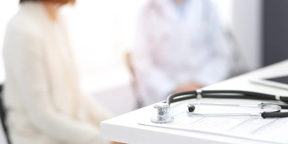 stethoscope, clipboard with medical form lying on hospital reception desk with laptop computer and busy doctor and patient communicating at the background medical tools at doctor working tablemedicine concept