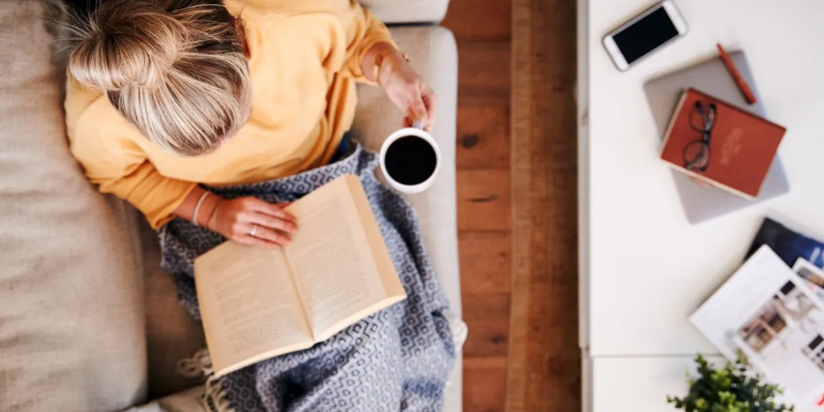 overhead shot looking down on woman at home lying on reading book and drinking coffee