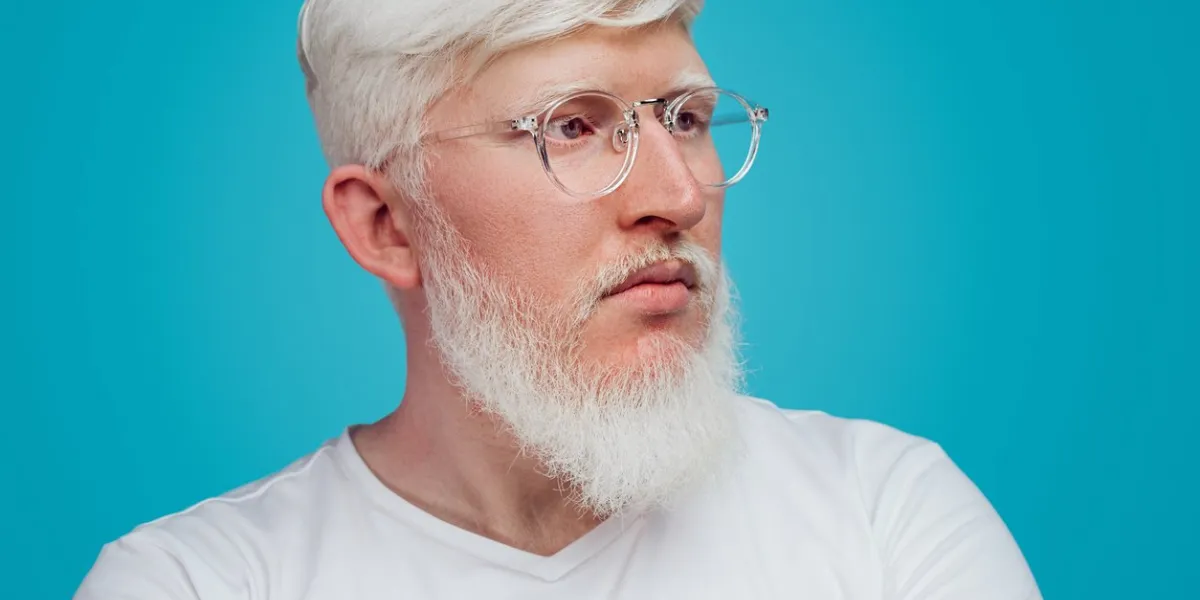 crop tranquil albino bearded male in glasses and casual white shirt looking away and contemplating against blue background in studio