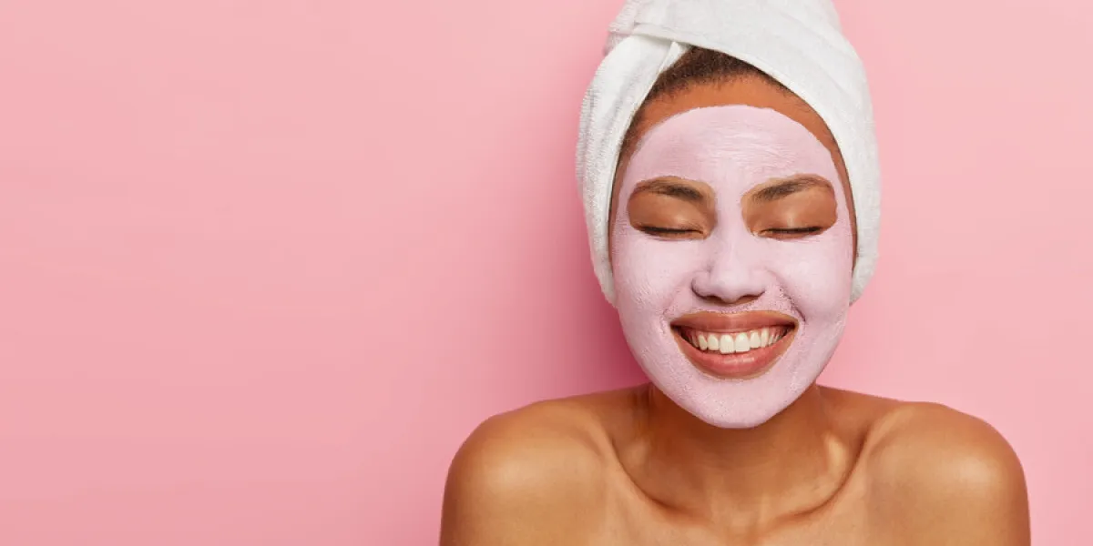 close up portrait of young female model applies homemade facial clay mask, has white towel wrapped around head, keeps eyes shut, smiles happily, models against pink background beauty treatment