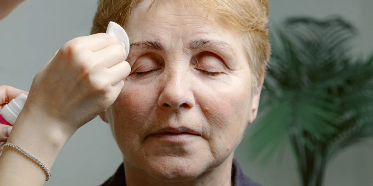one young caucasian young unrecognizable girl cosmetologist carefully removes the remaining paint from the right eyebrow of an elderly woman with closed eyes with a cotton pad, using a cleanser, in a home beauty salon, close-up side view