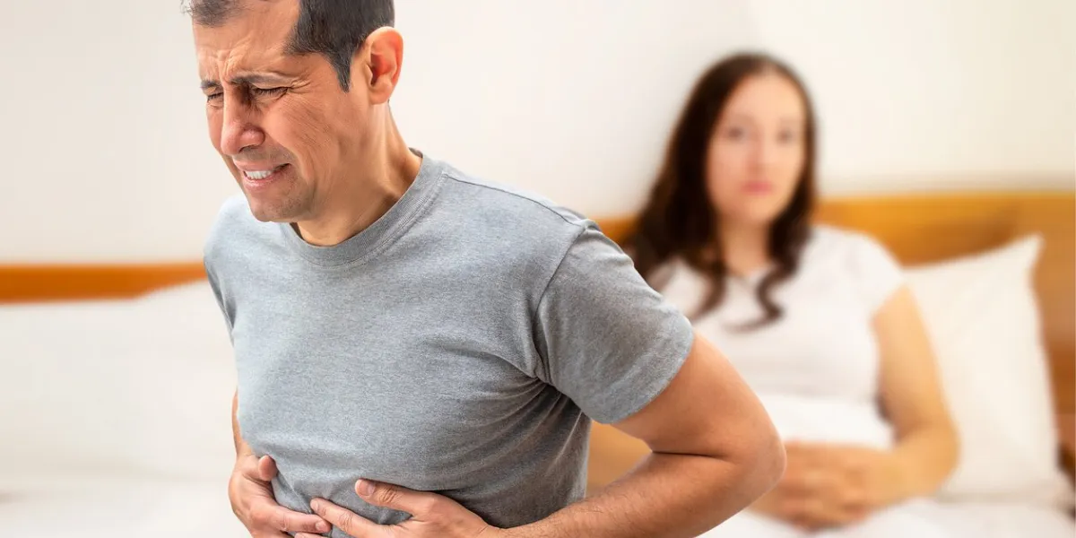 man suffering from stomach cramps sitting on his bed next to his wife
